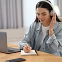 young woman with headphones taking notes at a table while listening to the lecture