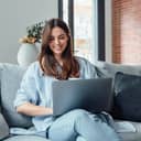 A concentrated millennial girl sitting on a couch working on the laptop, browsing internet and smiling.