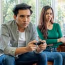 Man and woman sitting on couch with man holding a PlayStation controller and his partner looking at him disapprovingly