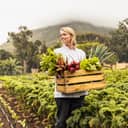 young female chef smiling while holding a crate of fresh vegetables while standing in an organic farm field