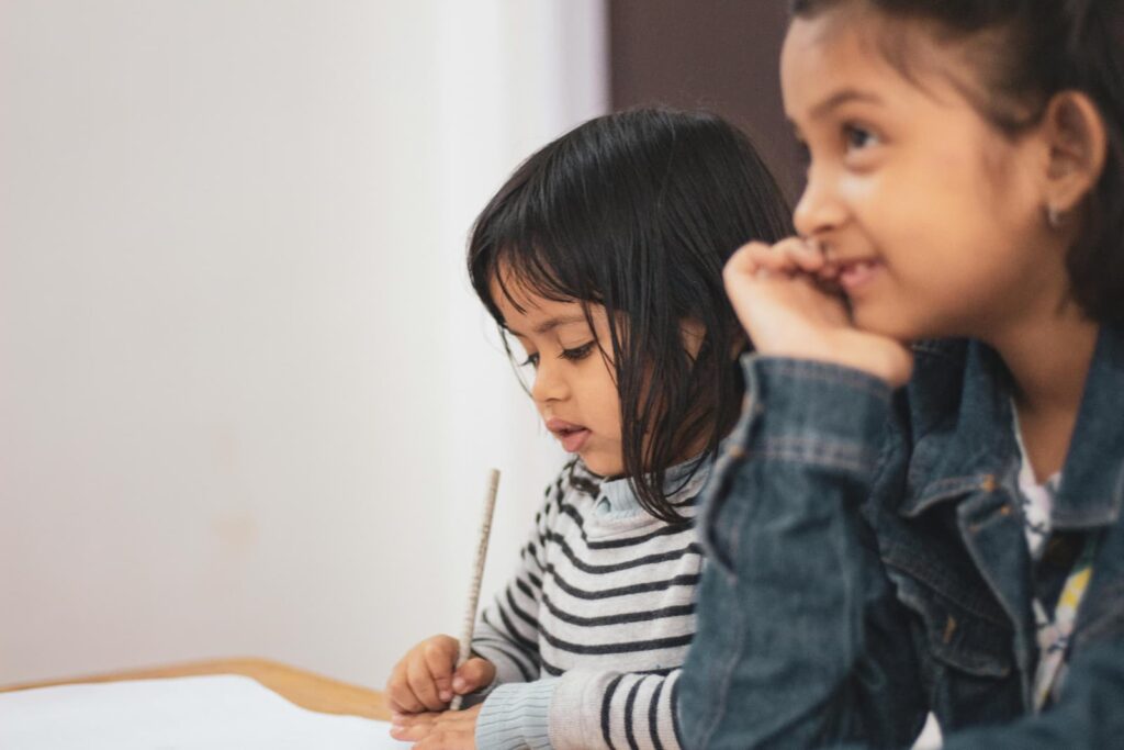 A little girl writing on a piece of paper next to an older, smiling girl
