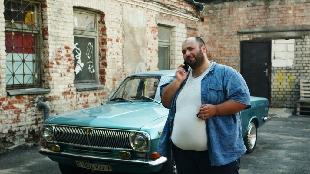 A heavyset man talking on a phone while standing beside a car outdoors