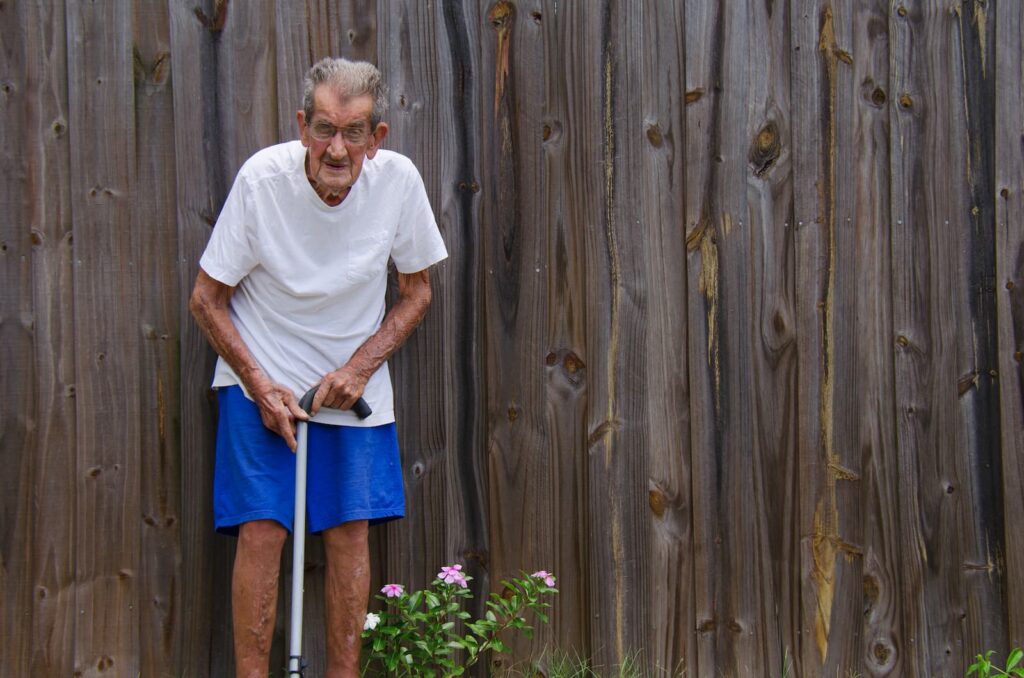 A frail, elderly man standing with a cane in front of a wooden gate outdoors