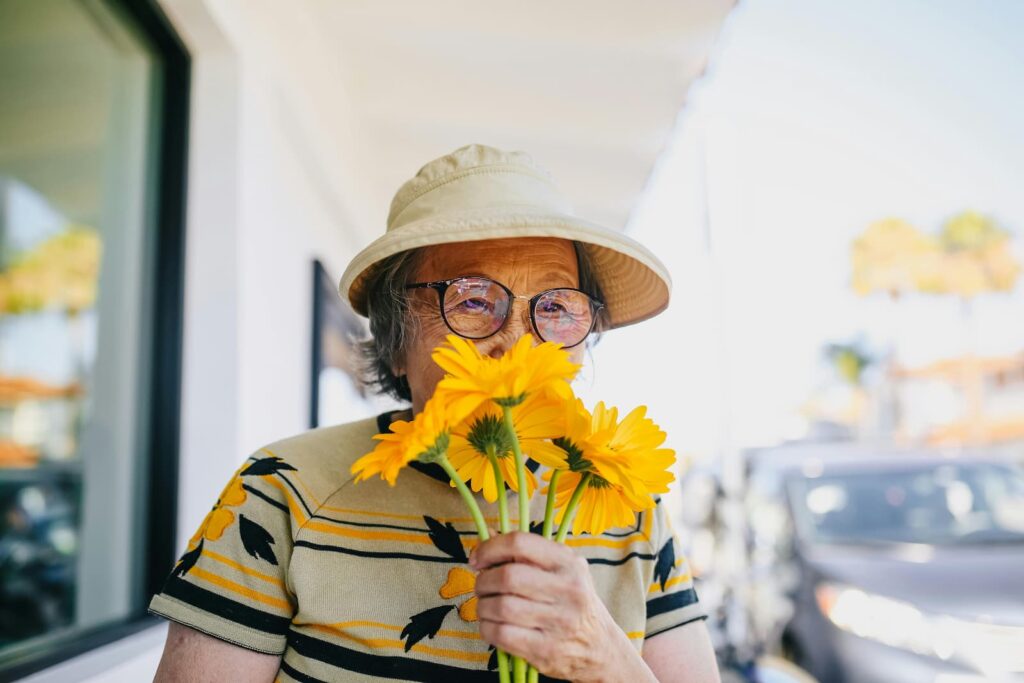 An elderly woman holding large, yellow flowers up to her nose