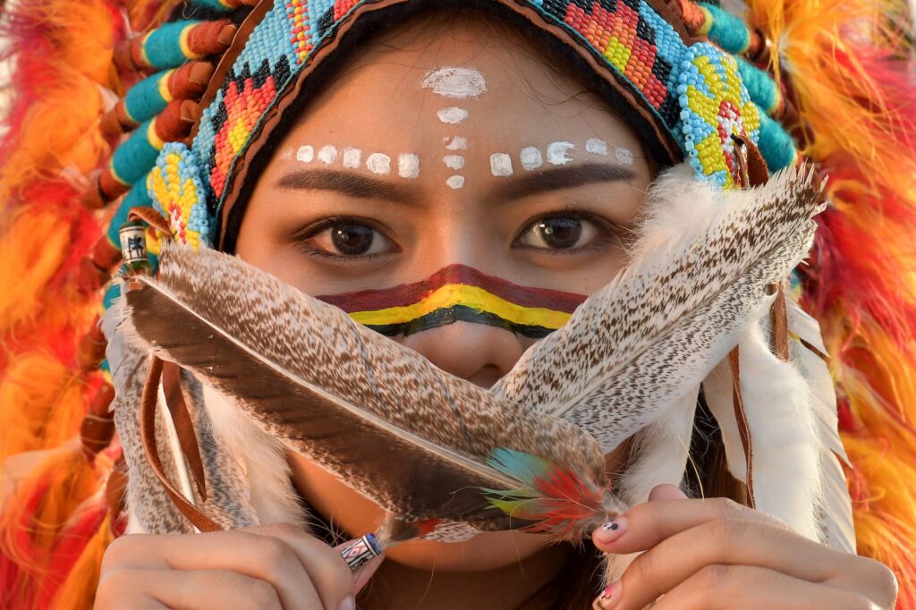 A young Native American woman in a traditional headdress holding two feathers in front of her face