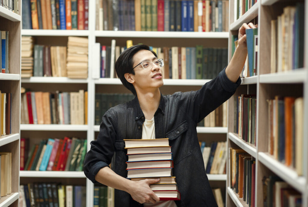 A young man holding a stack of books while walking through a library