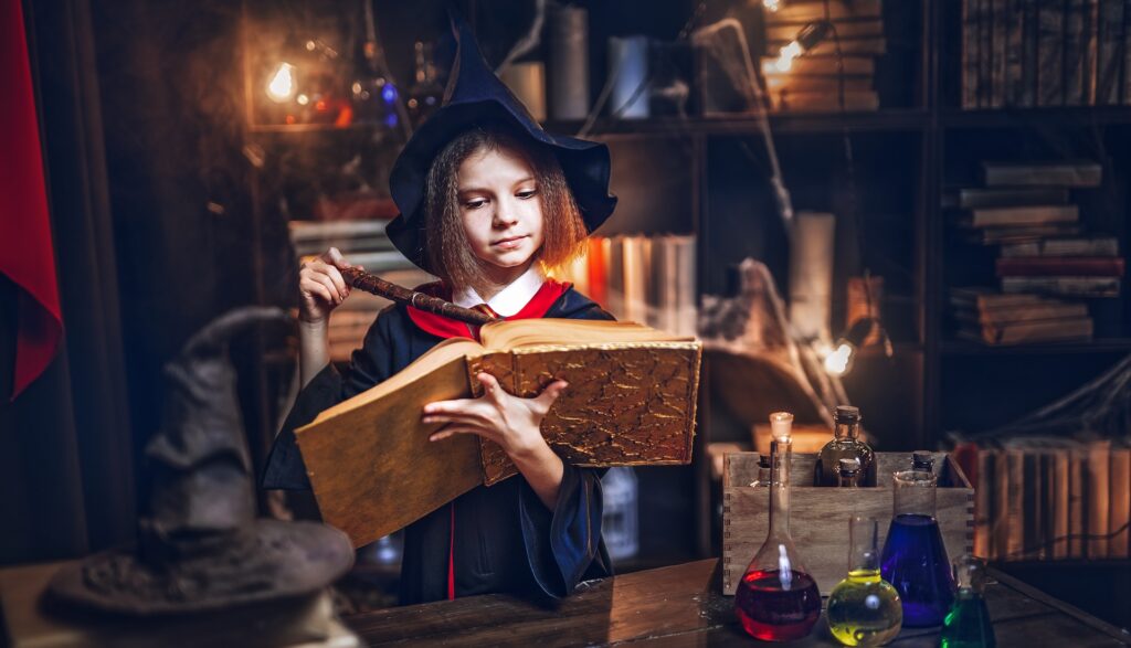 A little girl in a wizard robe and hat reading magic spells from an old book