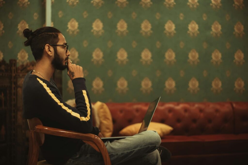 A seated man with a laptop holding his chin while thinking