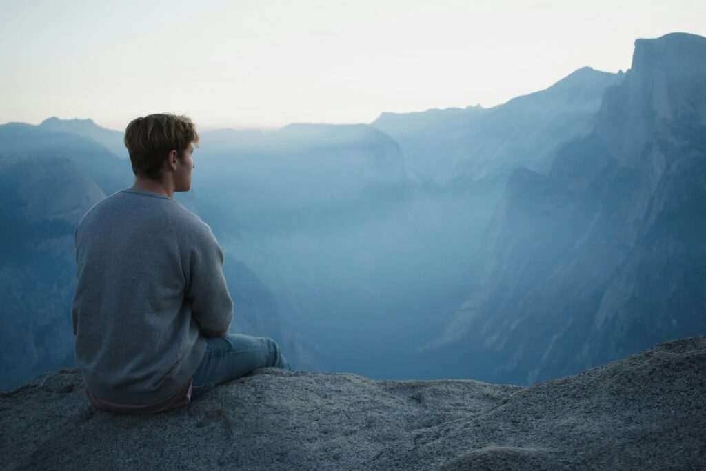 A man sitting on large rock, thinking while looking at the mountains in the distance